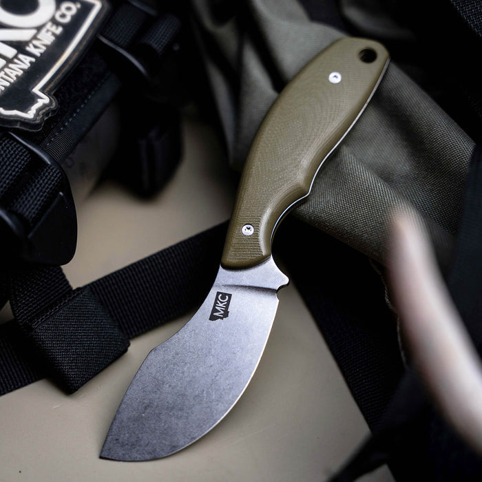 Olive PACKOUT SKINNER knife with a curved blade and textured handle, resting on tactical gear. Ideal for outdoor use.