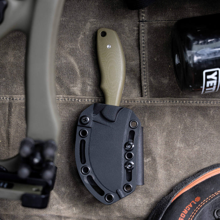 Olive green PACKOUT SKINNER knife with a black sheath, resting on a textured surface alongside outdoor gear.