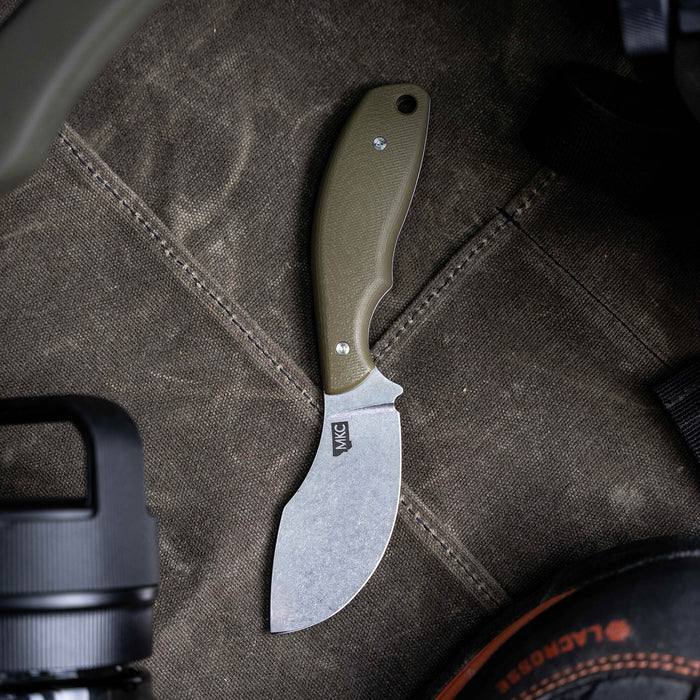 Olive green PACKOUT SKINNER knife with a curved blade, resting on a textured surface among outdoor gear.