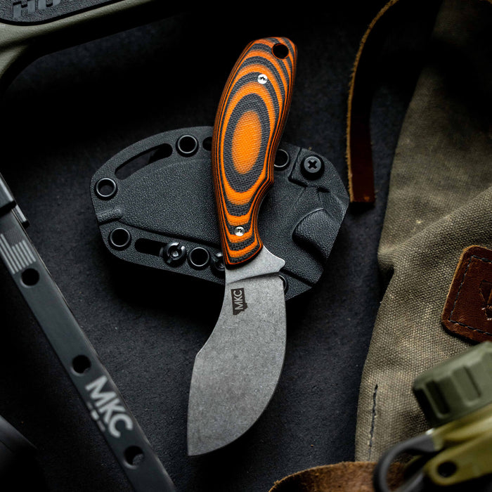 Orange and black PACKOUT SKINNER knife with a curved blade, resting on a black sheath and surrounded by tactical gear.