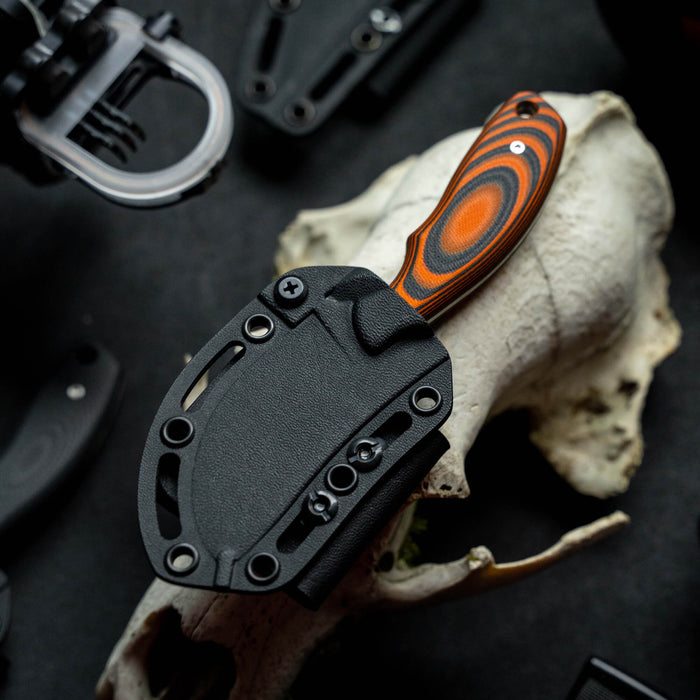 Orange and black PACKOUT SKINNER knife with a textured handle, displayed on a skull background, showcasing its rugged design.