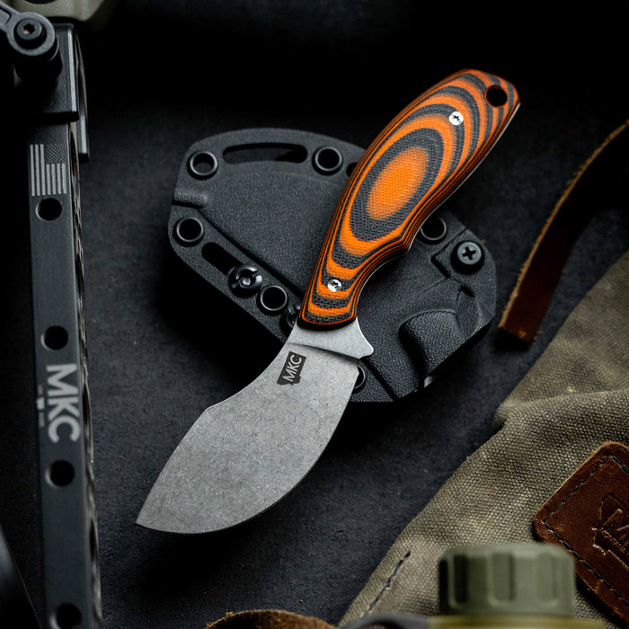 Orange and black PACKOUT SKINNER knife with a curved blade and ergonomic handle, displayed on a dark surface alongside gear.