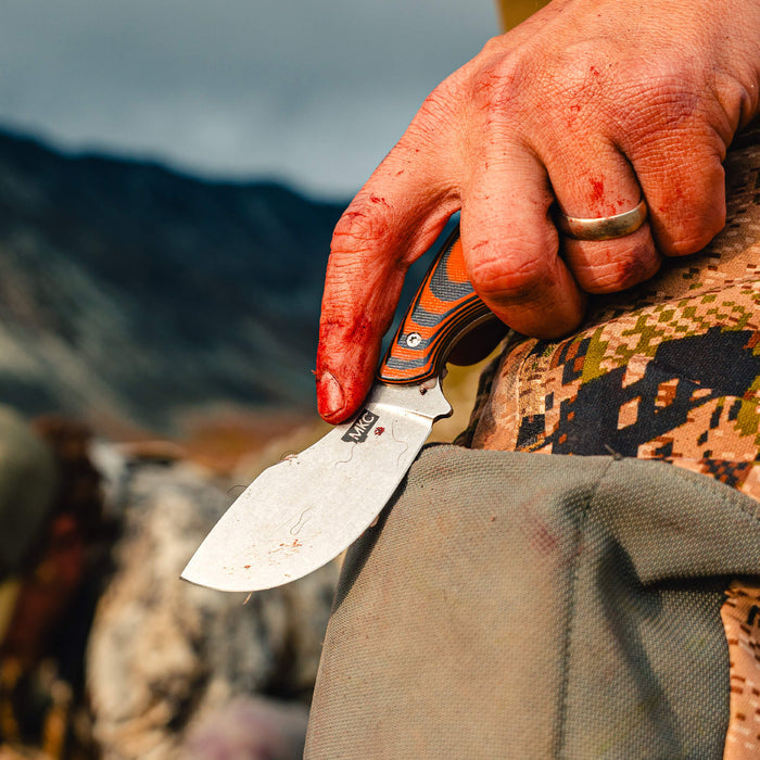 Close-up of a blood-stained hand holding the PACKOUT SKINNER knife with an orange and black handle, set against a rugged outdoor backdrop.
