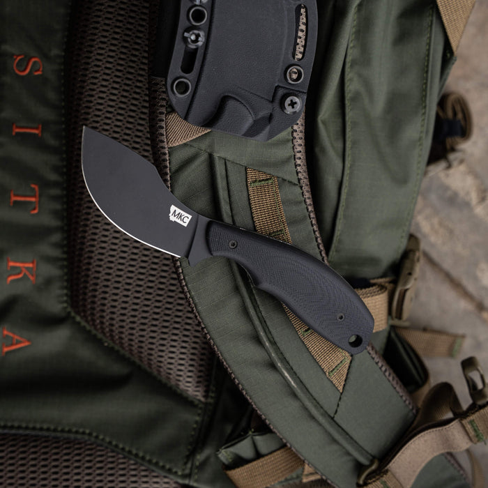 Montana Knife Company's PACKOUT SKINNER in PVD black, resting on a green tactical backpack, showcasing its sleek design.