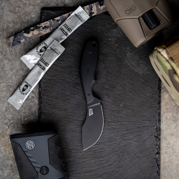 Montana Knife Company's PACKOUT SKINNER in PVD black finish, displayed on a slate surface with tactical gear.