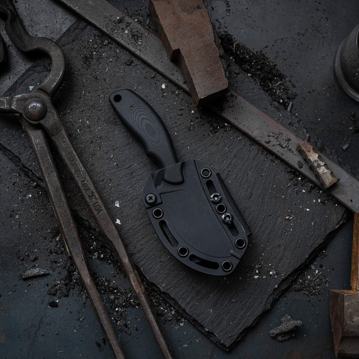 Black PACKOUT SKINNER knife with PVD finish on a textured slate surface surrounded by tools and debris.