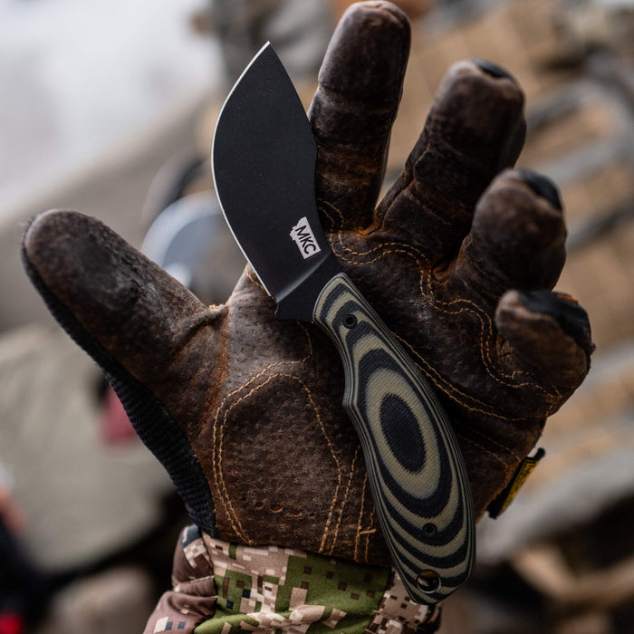 Montana Knife Company's PACKOUT SKINNER in green and black, held in a gloved hand, showcasing its curved blade design.
