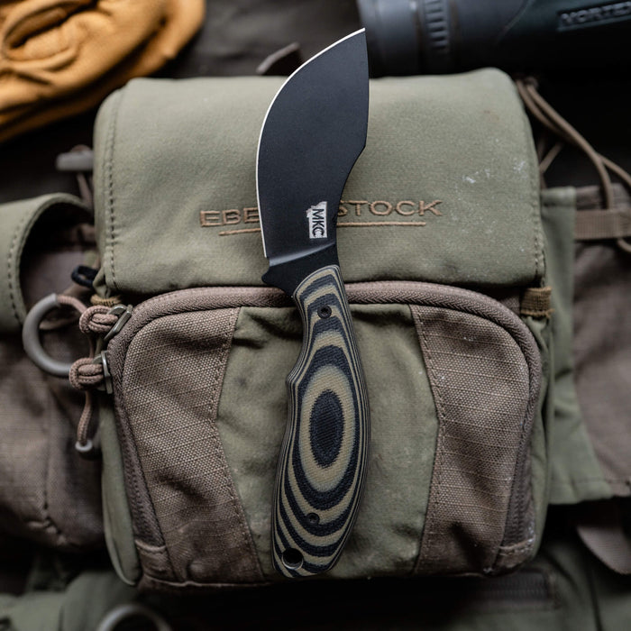 Montana Knife Company's PACKOUT SKINNER in green and black, featuring a curved blade and ergonomic handle, on a tactical pouch.