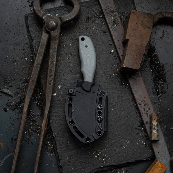 Grey PACKOUT SKINNER knife with PVD finish, displayed on a textured surface surrounded by tools and metal scraps.