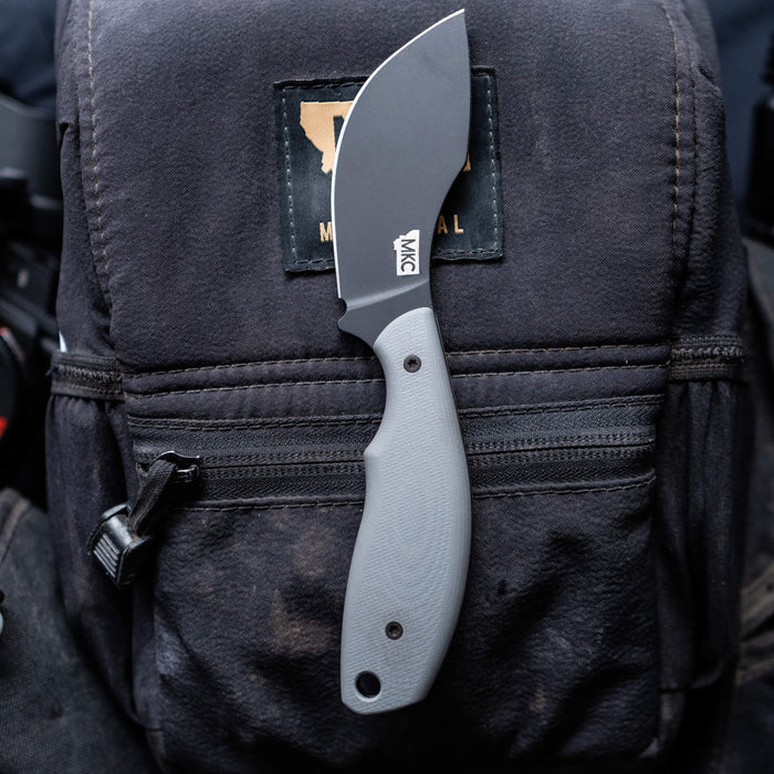 Montana Knife Company's PACKOUT SKINNER in grey, featuring a PVD-coated blade, rests on a rugged backpack.