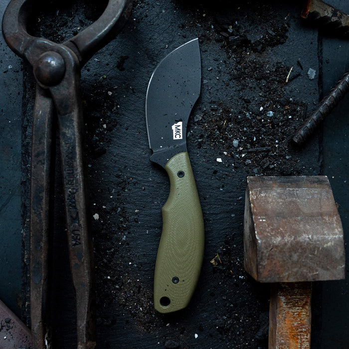 Montana Knife Company's PACKOUT SKINNER features a PVD-coated blade and olive green handle, set against a rugged workbench background.