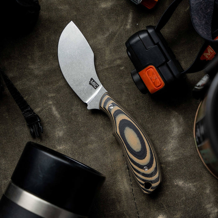 Tan and black PACKOUT SKINNER knife with a curved blade and ergonomic handle, surrounded by outdoor gear.