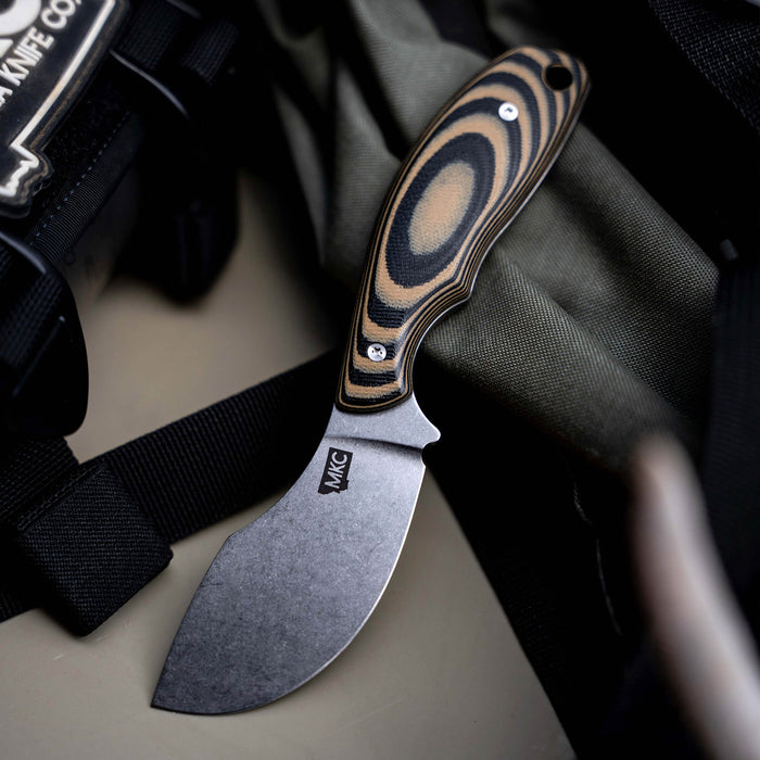 Montana Knife Company's PACKOUT SKINNER in tan and black features a curved blade and ergonomic handle, ideal for outdoor tasks.