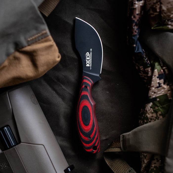 PACKOUT SKINNER PVD knife in red and black with a curved blade, resting on a dark fabric background among gear.