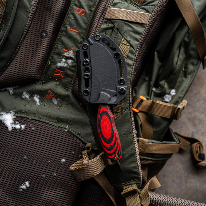 Red and black PACKOUT SKINNER PVD knife secured to a green outdoor backpack, showcasing its durable design for hunters.