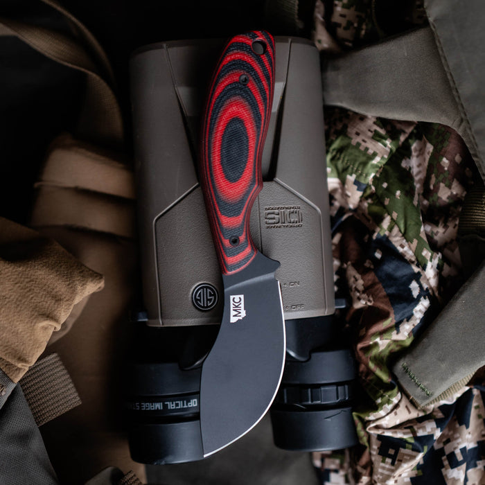 PACKOUT SKINNER PVD knife in red and black, resting on binoculars and camo fabric, showcasing its rugged design for outdoor use.