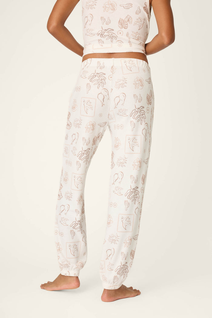 A person stands barefoot, shown from the back, wearing P.J. Salvage Palms Away Bistro Botanicals Banded Pants with a matching cropped tank top featuring abstract line art and plant patterns against a plain off-white background.