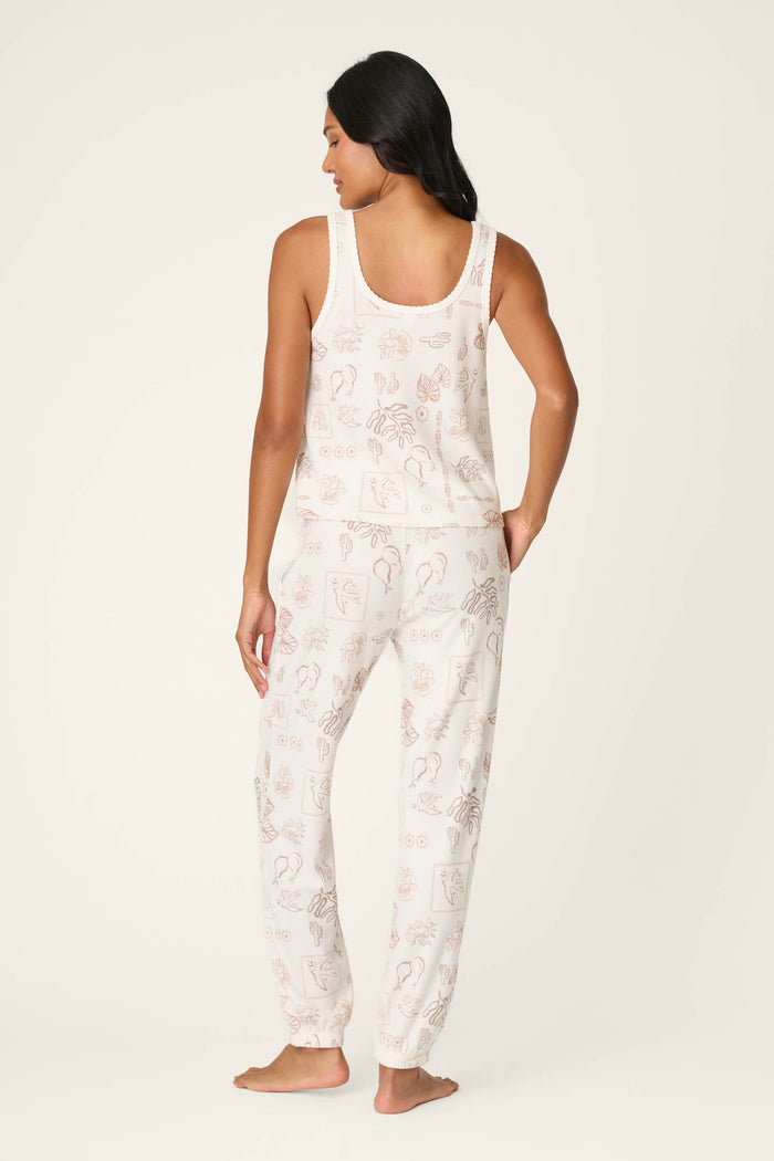 A woman with long dark hair stands barefoot, facing away, wearing the P.J. Salvage Palms Away Bistro Botanicals Long PJ Set—a sleeveless top and pants pajama set with a light cream background and subtle line art illustrations.