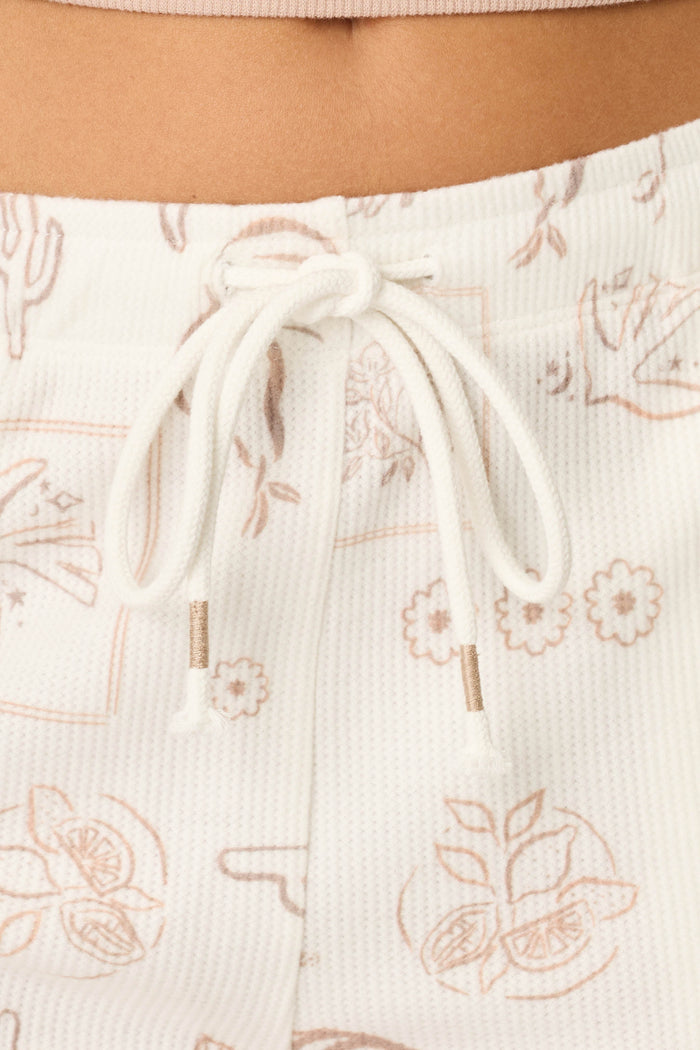 Close-up of a person's midsection wearing P.J. Salvage Palms Away Bistro Botanicals Shorts—white, textured with a drawstring and light brown botanical line art including flowers, citrus slices, and cacti. A beige top's waistband is visible above.