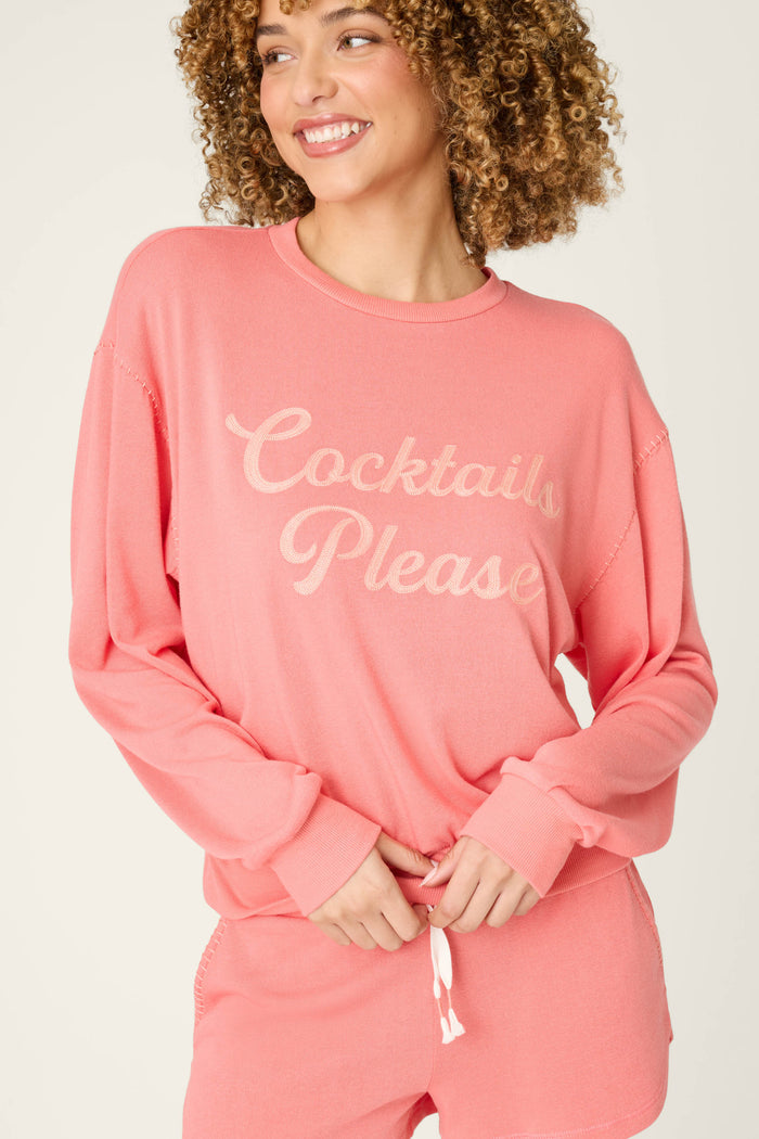 A person with curly hair wears the Paloma Mama Cocktails Please Graphic Sweatshirt by P.J. Salvage—a cozy pink French terry top with light script—while smiling and looking to the side, paired with matching shorts.