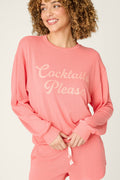 A person with curly hair wears the Paloma Mama Cocktails Please Graphic Sweatshirt by P.J. Salvage—a cozy pink French terry top with light script—while smiling and looking to the side, paired with matching shorts.