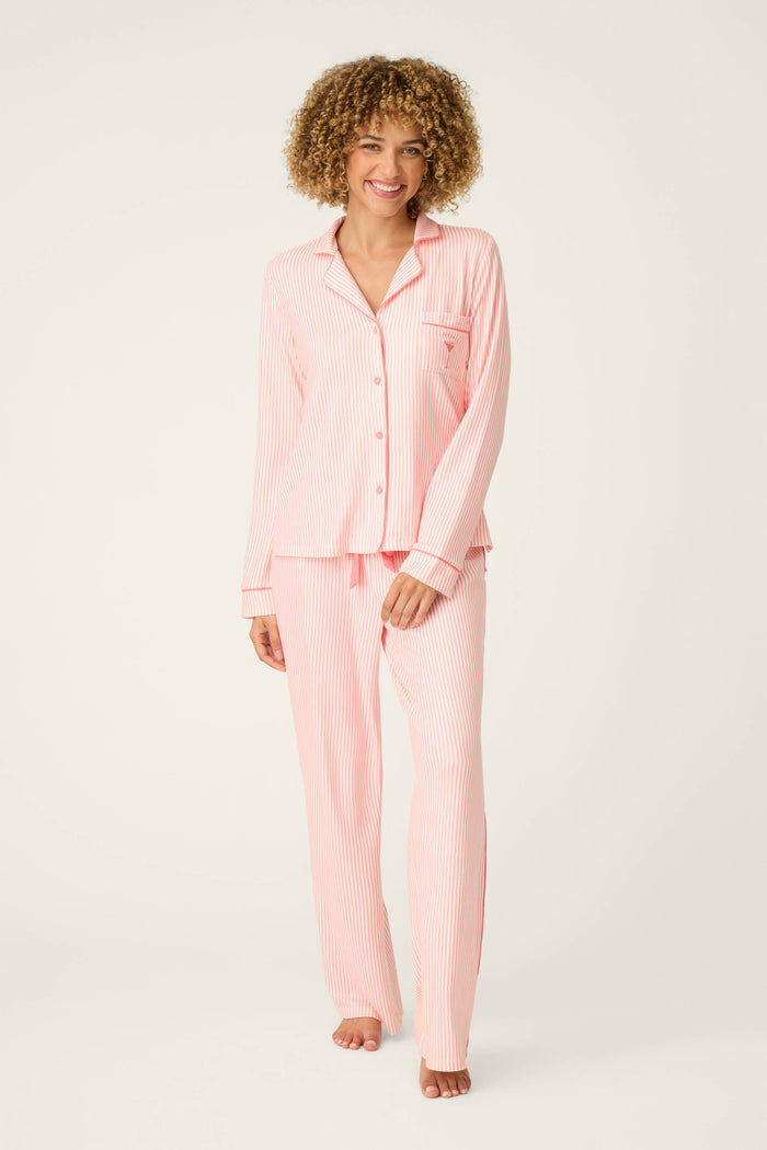 A person stands barefoot against a plain background, smiling in the Paloma Mama Sweet Stripes Long 3-Piece PJ Set by P.J. Salvage—a grapefruit pink striped sleepwear set with long sleeves, a collar, front buttons, and long pants.