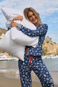 A smiling woman in P.J. Salvage’s Playful Prints Al Fresco Bistro Long PJ Set hugs a large white pillow on a beach, with hills and boats in the background under a partly cloudy sky.