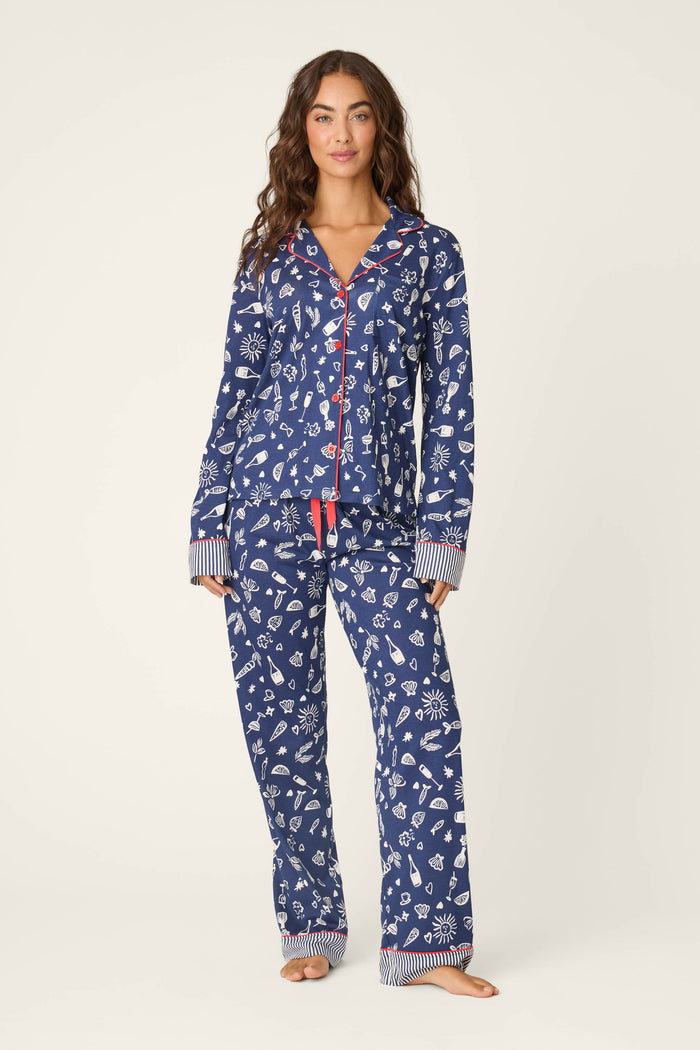 A woman stands barefoot against a plain light background, wearing the P.J. Salvage Playful Prints Al Fresco Bistro Long PJ Set—a blue cotton-modal jersey with white holiday prints, red buttons, and striped cuffs.