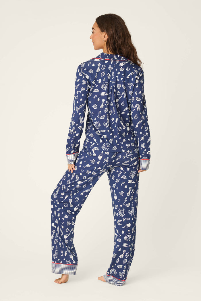 A woman stands barefoot, facing away, wearing the P.J. Salvage Playful Prints Al Fresco Bistro Long PJ Set featuring blue fabric with white winter-themed patterns and red-and-white striped cuffs on the sleeves and pants.