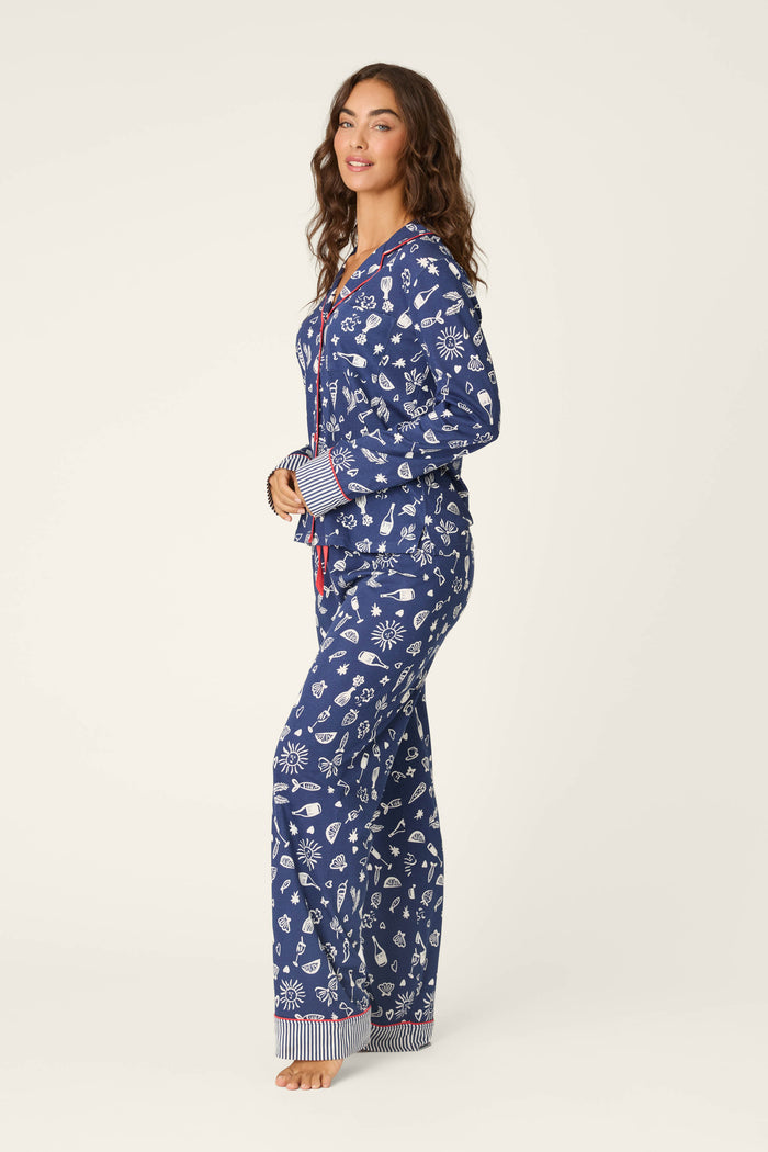 A woman stands barefoot in profile, wearing a P.J. Salvage Playful Prints Al Fresco Bistro Long PJ Set featuring blue fabric with white seashell and starfish designs, plus red-striped cuffs, set against a plain light background.