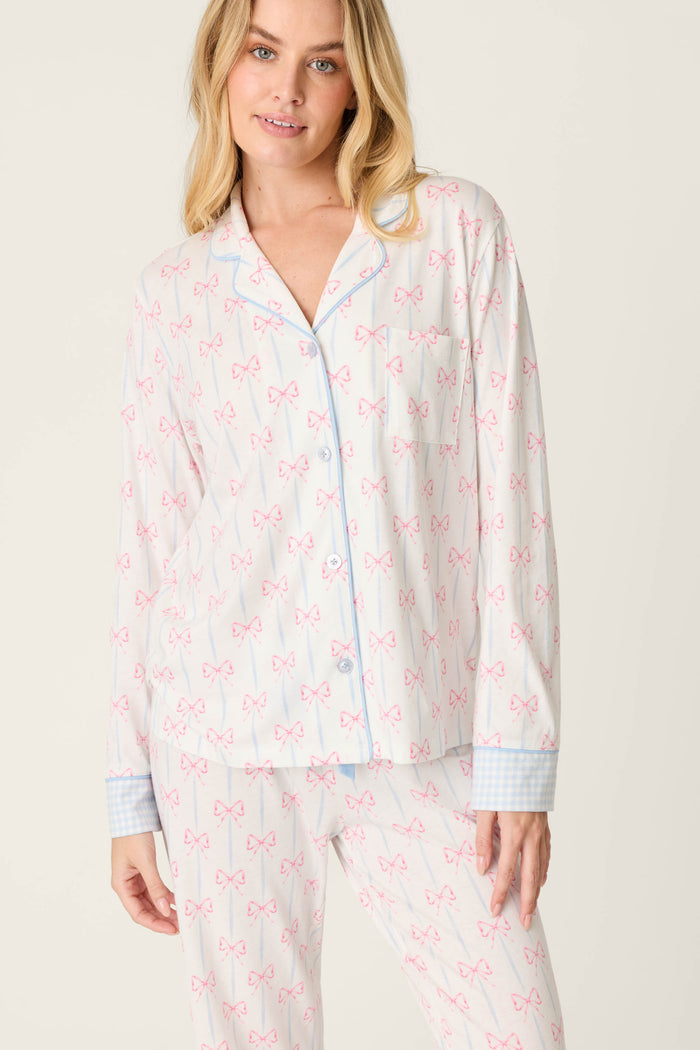 Wearing the Playful Prints All Wrapped Up Long Sleeve Top by P.J. Salvage, featuring soft cotton-modal jersey with a pink bow pattern, blue trim, and light blue checkered cuffs, a person stands against a plain light background.