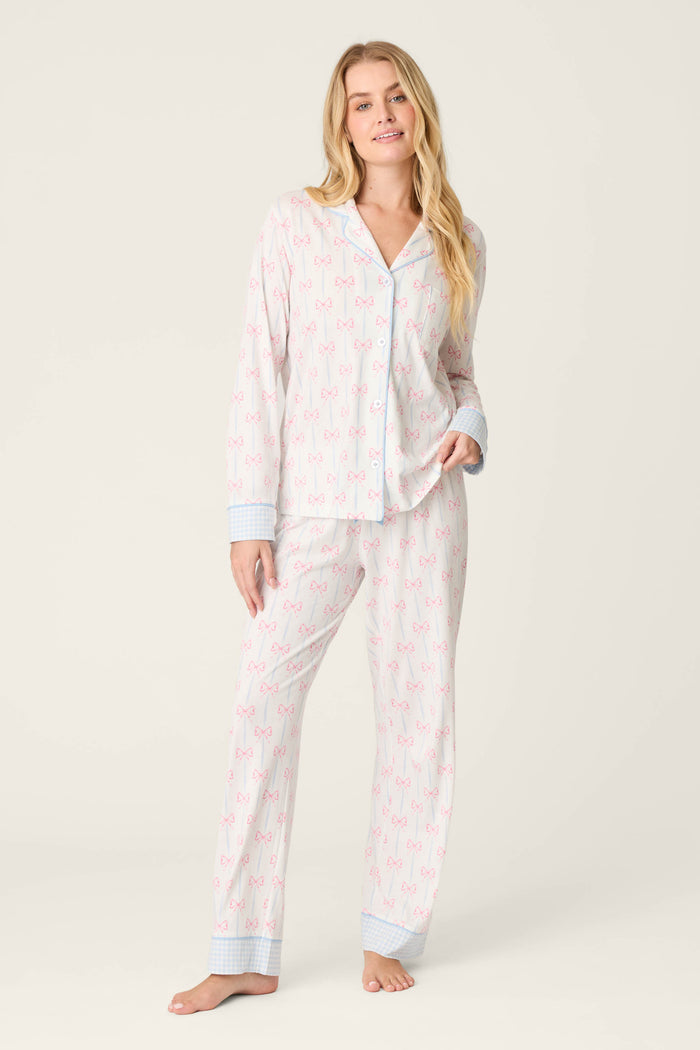 A woman stands barefoot in the P.J. Salvage Playful Prints All Wrapped Up Long Sleeve Top and matching pants, featuring pink bows and blue checkered cuffs, set against a plain, light background.