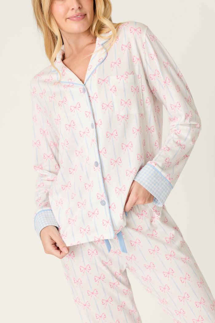 A woman wearing the Playful Prints All Wrapped Up Long Sleeve Top by P.J. Salvage, featuring pink bows and light blue checked cuffs and trim, stands against a plain light background.