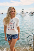 A smiling woman with long blonde hair wears the P.J. Salvage “Playful Prints Dolce Far Niente Short PJ Set” by the sea, with boats and a metal ladder visible in the background.
