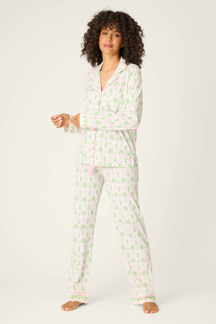 A person with curly hair stands barefoot, wearing the P.J. Salvage Playful Prints Island Time Long PJ Set in a light pink and green pattern on white, made from soft cotton-modal jersey, against an off-white background.
