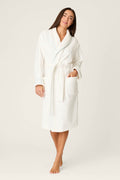 A woman stands barefoot in the P.J. Salvage Playful Terry Happy Hour Robe, a soft cotton terry robe with light blue trim, belt, and patch pockets. She appears relaxed against a plain light background, her long dark hair flowing.