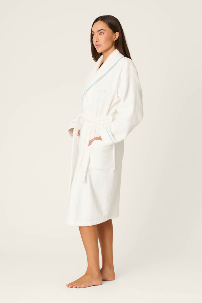 A woman stands barefoot on a light background wearing the P.J. Salvage Playful Terry Happy Hour Robe, a white terry style with light blue trim and waist tie. Her hands are in the robe’s pockets, and she looks forward with a neutral expression.