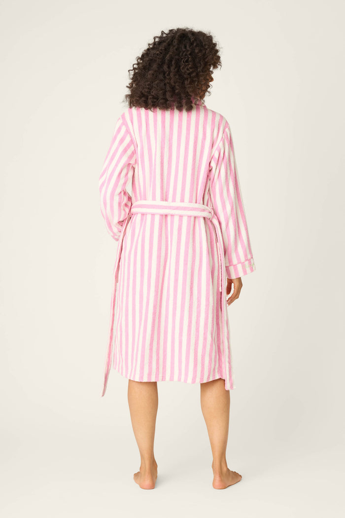 A person with curly hair stands barefoot facing away, wearing the P.J. Salvage Playful Terry Out Of Office Robe—a pink-and-white striped, knee-length style with a waist tie and soft cotton terry fabric—against a light plain background.