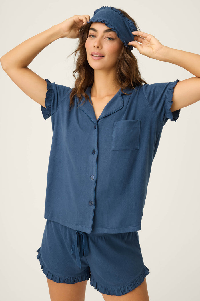 A woman in the P.J. Salvage Pointelle Essentials Deep Sea 3-Piece Shorts Set adjusts a blue sleep mask, standing against a plain background—a chic example of women’s sleepwear.