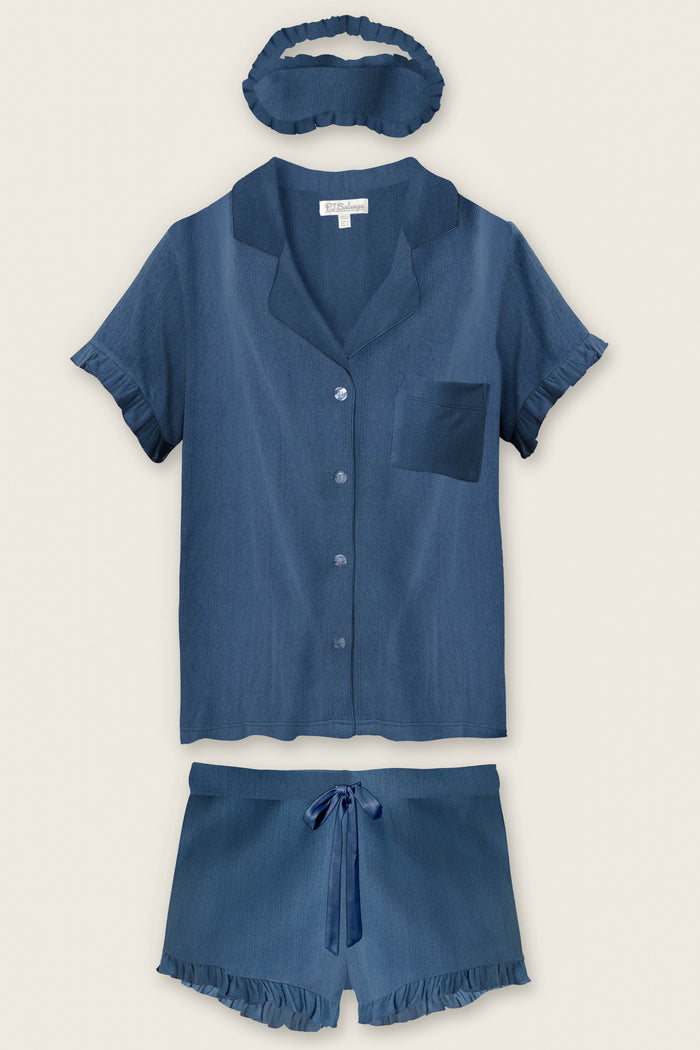 The P.J. Salvage Pointelle Essentials Deep Sea 3-Piece Shorts Set features a soft blue women’s sleepwear set with a short-sleeve button-up top, ruffled drawstring shorts, and a matching ruffled sleep mask laid flat on a neutral background.