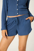 Wearing P.J. Salvage’s Pointelle Essentials Deep Sea Shorts with a light blue long-sleeve button-up top, one hand in their pocket and a hint of midriff showing.