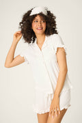 A woman with curly hair smiles while wearing the P.J. Salvage Pointelle Essentials Ivory 3-Piece Shorts Set and a white sleep mask, standing against a plain background and looking slightly to the side.