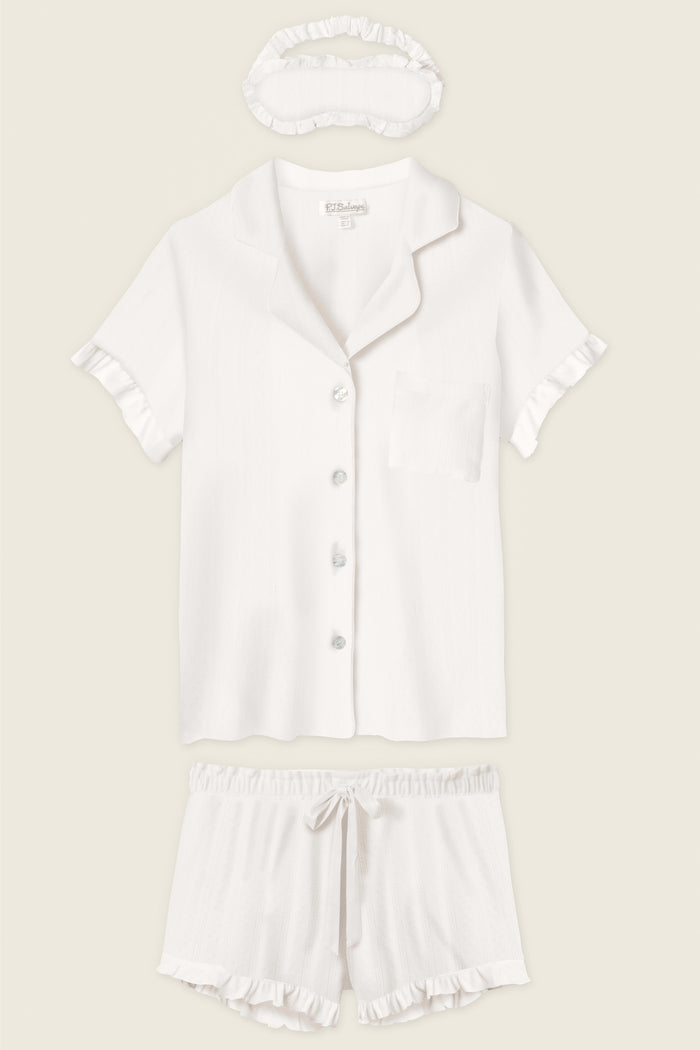 The P.J. Salvage Pointelle Essentials Ivory 3-Piece Shorts Set includes a short-sleeve button-up shirt with chest pocket, matching drawstring shorts, and a sleep mask—all in white with ruffle trim.