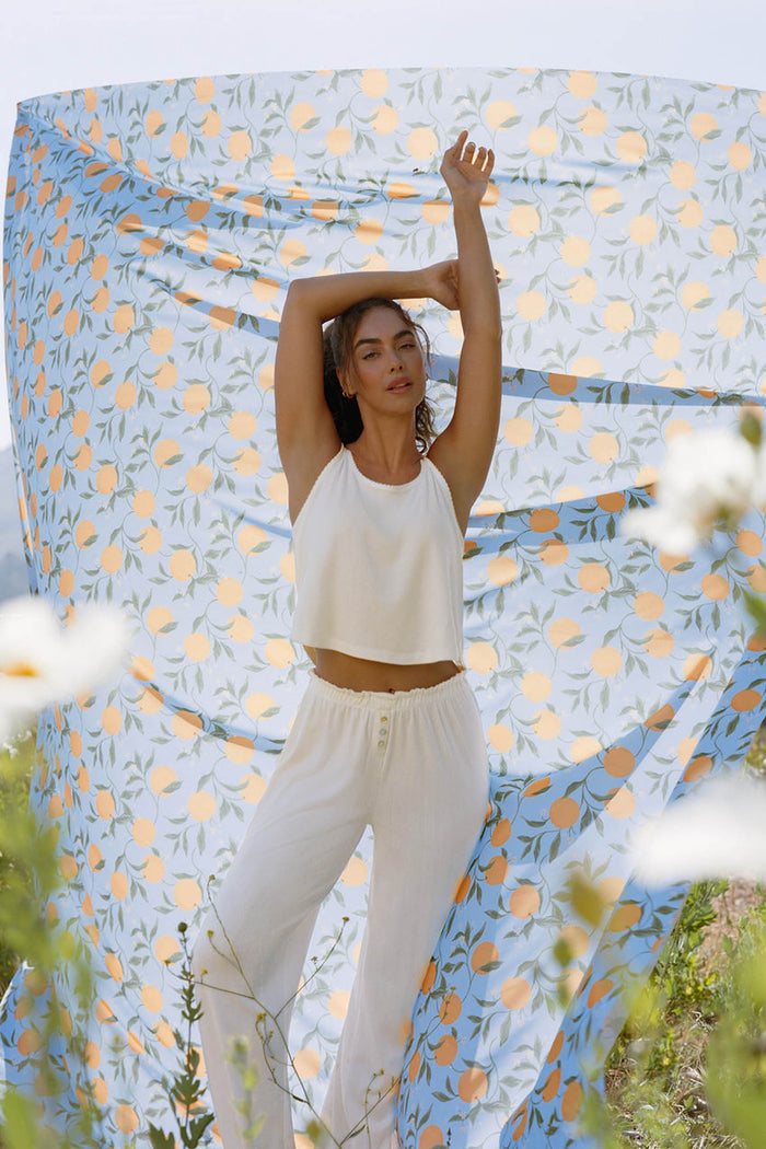 A woman stands outdoors with arms raised, wearing the P.J. Salvage Pointelle Essentials Ivory PJ Set. Behind her is a lemon and leaf print fabric, with flowers and greenery visible in the foreground.