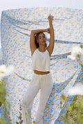 A woman stands outdoors with arms raised, wearing the P.J. Salvage Pointelle Essentials Ivory PJ Set. Behind her is a lemon and leaf print fabric, with flowers and greenery visible in the foreground.
