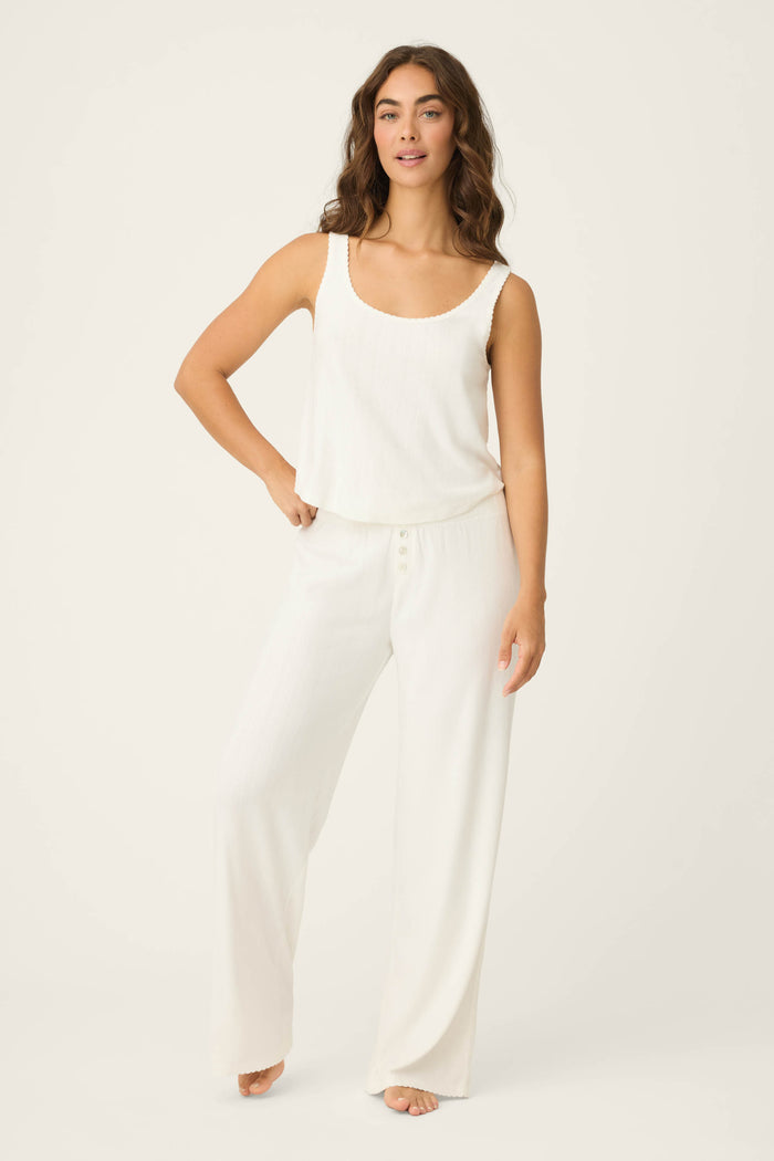 A woman stands barefoot against a plain light background, modeling the P.J. Salvage Pointelle Essentials Ivory PJ Set: a sleeveless white tank top with loose white pants. One hand is on her hip, the other rests by her side.