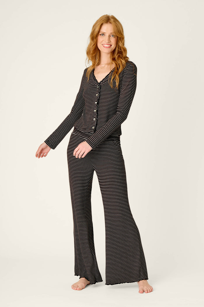 A person with long red hair smiles while wearing the Pretty Stripes Onyx Pointelle Cardigan by P.J. Salvage, standing barefoot against a plain light background.