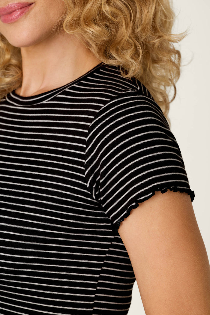 A woman with curly blond hair is shown from the shoulders up against a light background, wearing the Pretty Stripes Onyx Pointelle Short Sleeve Top by P.J. Salvage—a black and white striped top featuring ruffled edges on the sleeves.
