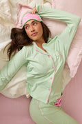 A person lounges on a bed in the P.J. Salvage Pupcation Palms Kiwi Stripe 3-Piece PJ Set, featuring green stripes and pink trim, paired with an embroidered sleep mask. The bedding is white and pale pink, with an arm resting above their head.