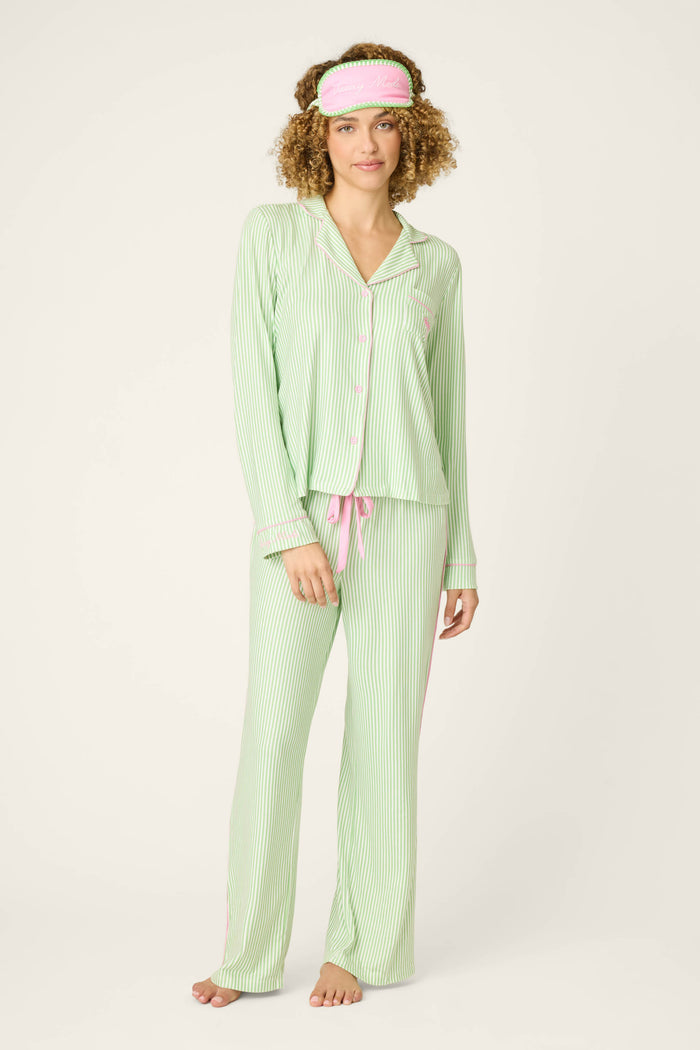 A woman stands barefoot facing forward in the P.J. Salvage Pupcation Palms Kiwi Stripe 3-Piece PJ Set, featuring green and white stripes with pink accents and a matching sleep mask, against a plain light background.